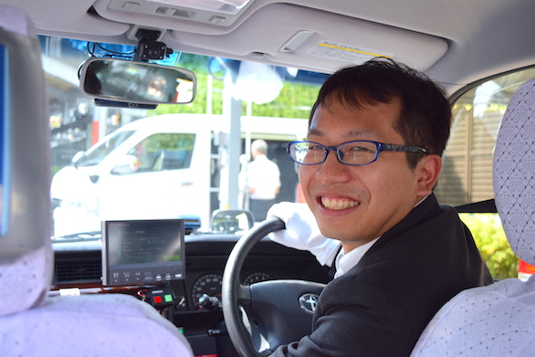 タクシーの運転席から笑顔で振り返る国際自動車（kmタクシー）の男性タクシードライバー青島さんの写真
