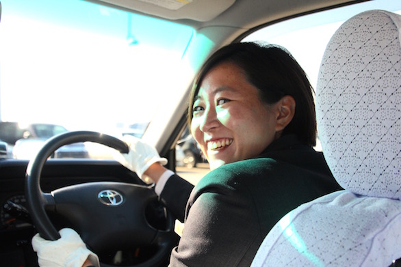 タクシーの運転席から笑顔で振り返る、国際自動車（㎞タクシー）株式会社に第二新卒で入社した女性タクシードライバー河原さんの写真