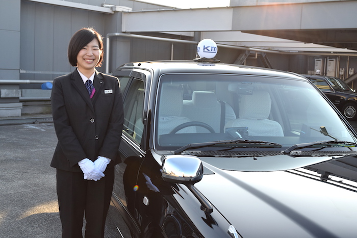 タクシーの横に笑顔で立っている、国際自動車（kmタクシー）の女性タクシードライバーで、新卒女性班長の成毛さんの写真