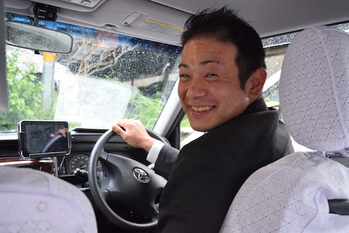 国際自動車（㎞タクシー）横浜のタクシードライバー瓜生さんが運転席から笑顔で振り返っている写真