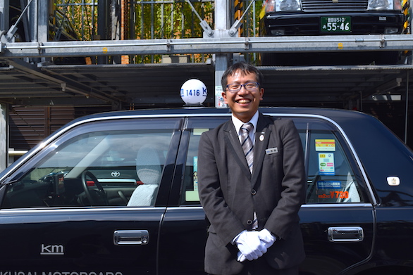 タクシーを背に笑顔で立っている国際自動車（kmタクシー）の男性タクシードライバー青島さんの写真