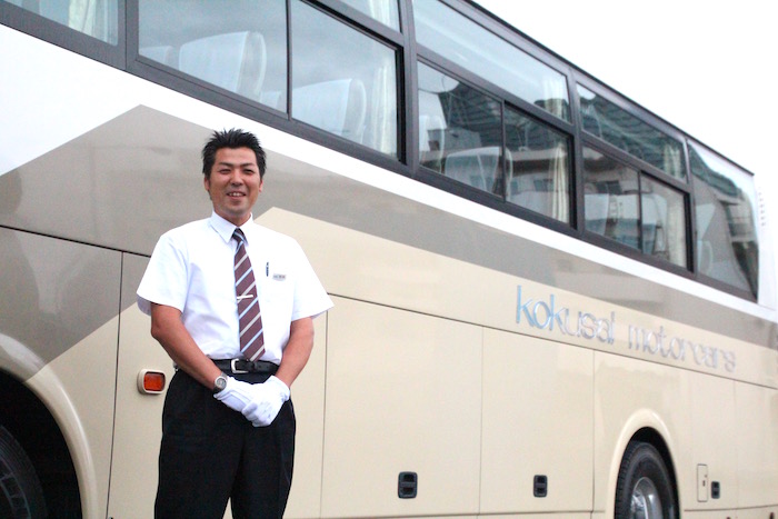 観光バスを背に笑顔で立っている、ケイエム観光バス株式会社のバスドライバー阿部さんの写真