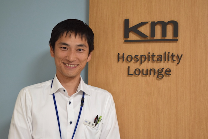 「km Hospitality Lounge」の文字が掲げられた壁の前に笑顔で立っている、国際自動車（㎞タクシー）の元タクシードライバーで現在は人財採用課（人事）の福島さんの写真